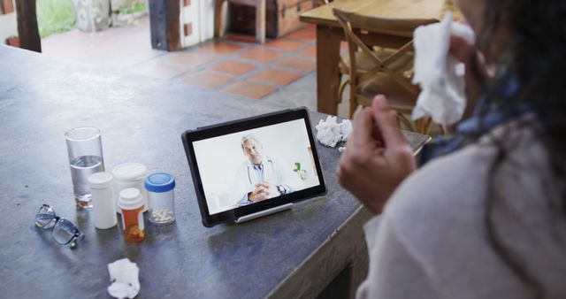 Individual sitting at table having virtual consultation with doctor on tablet, medications and tissues nearby. Useful for content on telemedicine, online healthcare services, and home-based medical care solutions.