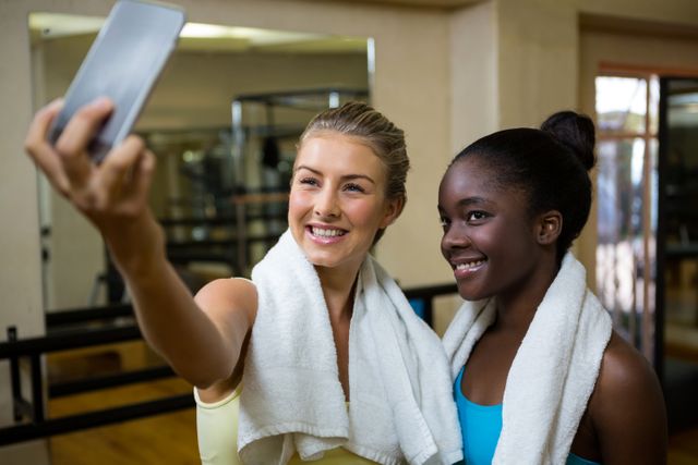Two fit women are taking a selfie with a mobile phone after a workout in a gym. Both are smiling and have towels around their necks, indicating they have just finished exercising. This image can be used for promoting fitness, healthy lifestyles, gym memberships, or social media content related to exercise and wellness.
