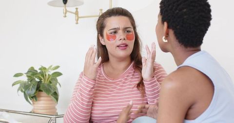 African american friends applying under-eye gel patches, talking, gesturing on sofa