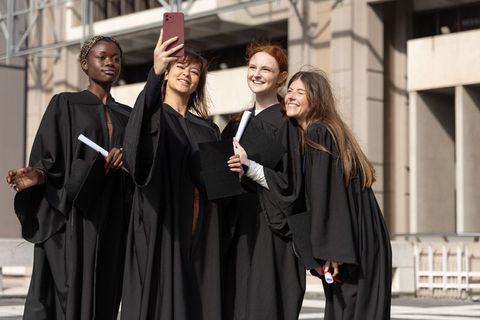 Diverse graduates taking selfie on campus in black gowns celebrating diploma achievement
