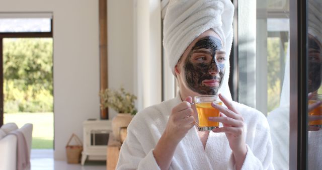 Woman standing near window dressed in white bathrobe and towel on head, enjoying herbal tea. She is wearing a black face mask, emphasizing peaceful, relaxed mood. Perfect for content related to self-care routines, skincare products, wellness, and home relaxation.