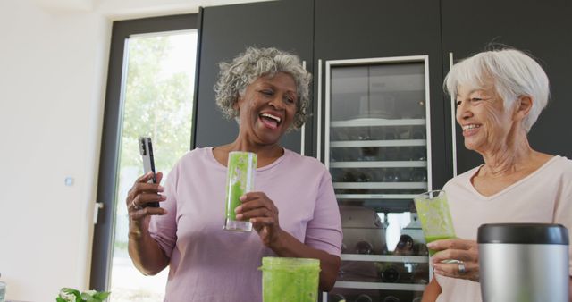 Two senior women smiling and enjoying green smoothies in modern, stylish kitchen. They appear happy and engaged, making this image perfect for promotional content related to healthy living, senior wellness, social activities for seniors, and modern kitchen design.