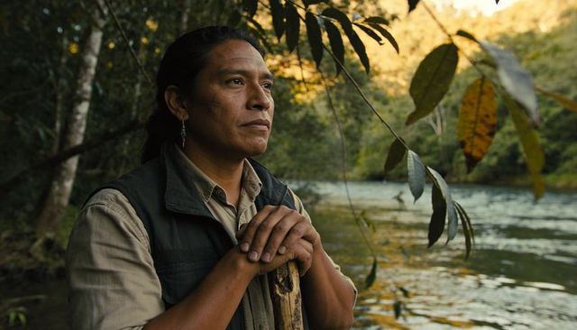 Native american outdoorsman leaning on wooden staff gazing over river at golden hour