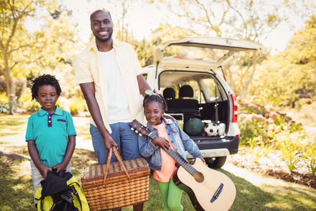 African American father and two children enjoying a sunny day at the park. The father holds a picnic basket while the daughter holds a guitar, and the son stands beside them. Ideal for use in advertisements, family-oriented content, and articles about outdoor activities, family bonding, and leisure time.