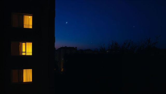 Apartment windows glowing at dusk with silhouette watching crescent moon over quiet city