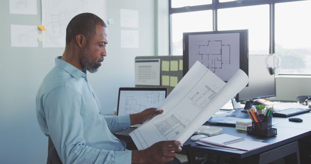 Architect in modern office reviewing blueprints at desk with computer screens displaying design plans. Used for concepts related to architecture, engineering, project management, professional work environments, and creative design. Suitable for illustrating office settings, technical work, or team-based projects.