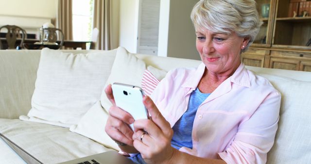 Senior woman sitting on couch smiling while using smartphone, indicating comfort with technology. Perfect for depicting senior lifestyle, technology adoption among elderly, and home comfort. Could be used in advertising for mobile devices, eldercare technology services, or lifestyle blogs about aging.