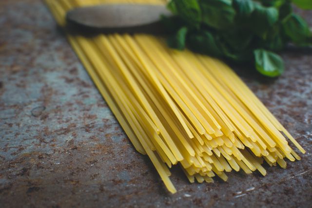 Close-up of uncooked spaghetti pasta on rustic surface accompanying fresh basil leaves and wooden spoon. Perfect for illustrating recipes, gourmet cooking blogs, Italian cuisine articles, food preparation concepts and kitchen essentials.