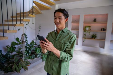 Young asian man texting on smartphone in modern home with floating stairs and plants