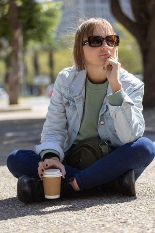 Young woman sitting cross-legged holding coffee cup wearing denim jacket and sunglasses