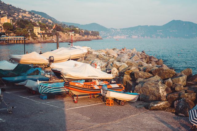 Boats are docked on a rocky shoreline in an Italian coastal village during sunset. Scene captures the tranquil beauty of the Mediterranean coast, highlighting small boats covered and resting on the dock, along with a rocky breakwater extending into calm waters. Ideal for travel brochures, tourism ads, or websites promoting coastal getaways and scenic coastal views.
