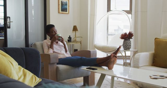 Happy african american woman sitting on armchair in living room, drinking coffee. domestic life, spending time at home.
