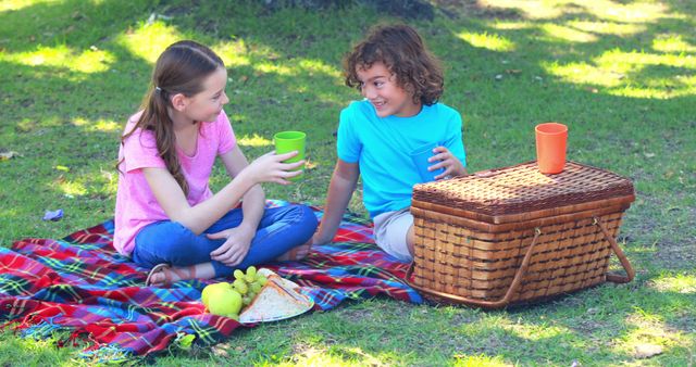 Two children, a Caucasian girl and a boy with curly hair, are enjoying a picnic on a sunny day, with copy space. They are sitting on a blanket with a basket and snacks, cheerfully toasting with their cups.