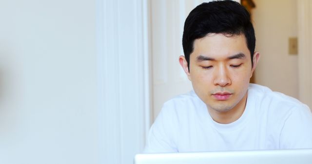 This image shows a young Asian man focused on his laptop screen while working from home. Ideal for illustrating concepts of remote work, freelancing, home offices, or concentration on tasks. Useful in blog posts, articles, advertisements, and websites related to technology, work-from-home setups, and productivity.