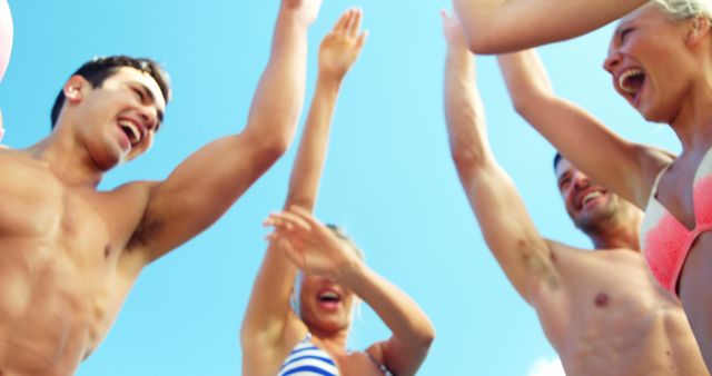 A group of joyful young adults is celebrating under a clear blue sky, with copy space. Their raised hands and bright smiles convey a sense of happiness and freedom during an outdoor gathering.