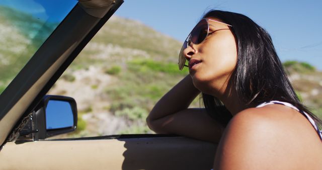Young woman enjoying a summer drive in a convertible car, wearing sunglasses, and relaxing with arm on car door. Ideal for use in travel advertisements, road trip promotions, lifestyle blogs, and content highlighting freedom, adventure, and outdoor activities.