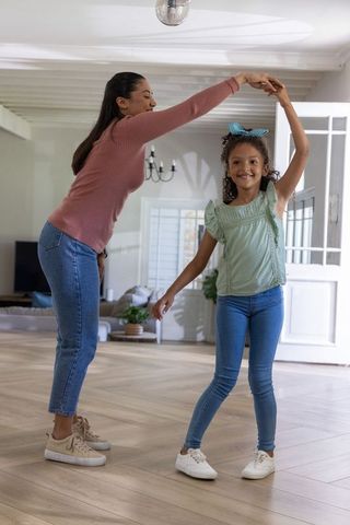 Diverse mother twirling daughter in bright living room, joyful family dance on hardwood