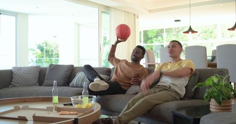 Friends watching game on sofa with basketball and pizza in bright casual home living room