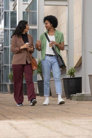 Diverse coworkers walking and talking while holding coffee in modern urban office plaza