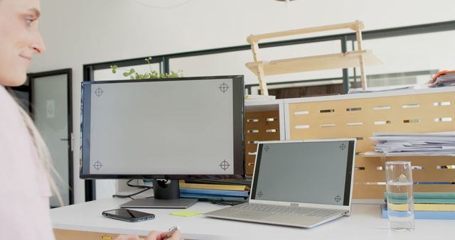 A woman working from a home office desk. She is looking at a computer monitor next to a laptop. The workspace includes stacks of papers, a smartphone, and a glass of water. Use for illustrating remote work setups, home office organization, and modern work-life balance.