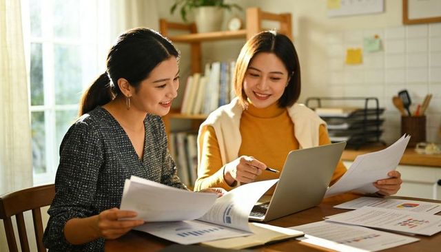 Two women collaborating on financial charts and laptop at bright kitchen workspace