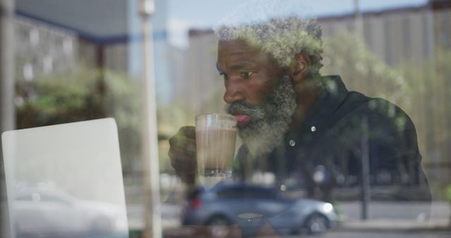 Senior man seen through window working on a laptop while drinking coffee. Reflections suggest a busy urban environment. This can be used for topics related to remote work, entrepreneurship, senior individuals in tech, or modern lifestyle themes.