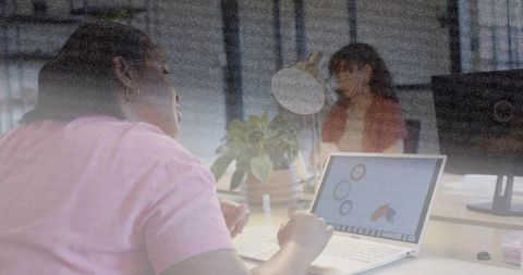 Typing woman in pink t-shirt working on laptop with analytics charts during team meeting
