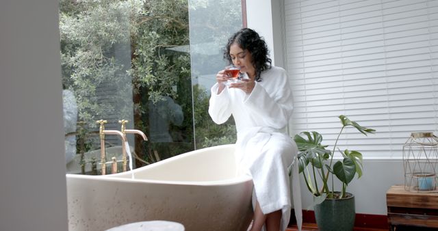 A woman wearing a bathrobe is sitting on the edge of a modern bathtub, enjoying a warm cup of tea. The bathroom features a large window with a view of lush greenery, adding to the serene and relaxing atmosphere. Indoor plants and minimalistic decor enhance the tranquil setting. This image can be used for promoting self-care, wellness, spa services, relaxation products, or bathroom design and decor.