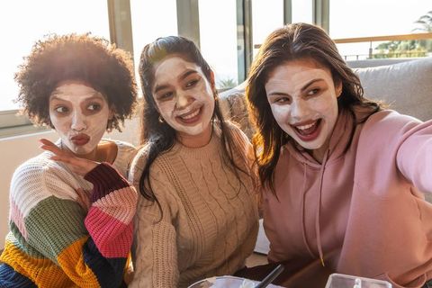 Multicultural friends enjoying cozy skincare night, wearing face masks and taking selfie