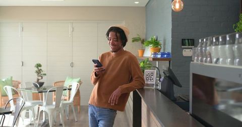 African american man leaning on cafe counter using smartphone, smiling in coffeehouse