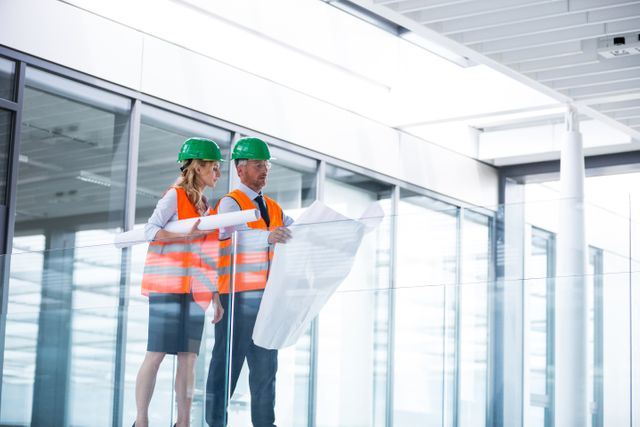 Architects in safety vests and hard hats are discussing a blueprint in a modern office corridor with glass walls. Ideal for use in articles or presentations about architecture, construction planning, teamwork in engineering, and professional collaboration in modern workspaces.