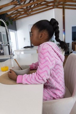 African american girl sitting at kitchen island wearing pink robe holding bowl and spoon