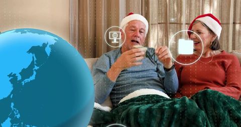 Senior couple wearing santa hats enjoying cozy tea on sofa with digital globe ui overlay