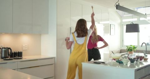 Female friends cooking and dancing at bright modern kitchen island with wooden spoon play