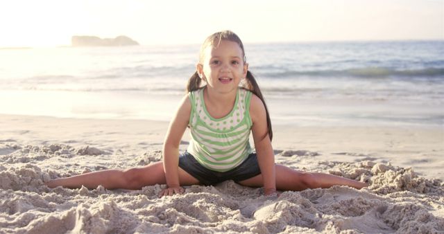 Young girl in green striped tank top and shorts doing splits on sandy beach at sunset. Ideal for use in campaigns promoting outdoor kids activities, healthy lifestyle, and summer leisure activities for children.