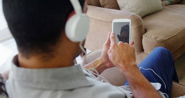 Man sitting on couch wearing headphones and using a tablet, shot from behind. Perfect for use in technology, leisure, and lifestyle content. Ideal for promoting headphones, tablets, and relaxation products or services.