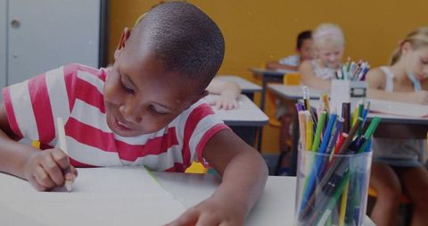 Writing boy focusing at classroom desk with pencil cup and colorful school supplies