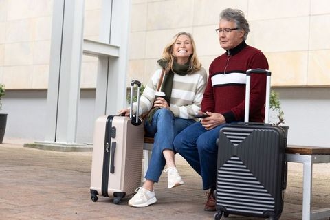 Mature couple smiling, waiting with suitcases and coffee and checking phone on urban bench