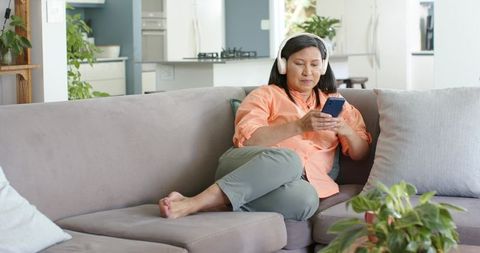 Relaxing mature asian woman listening with headphones and browsing smartphone on sofa