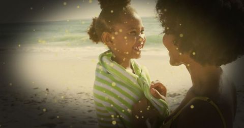 Smiling mother holding daughter wrapped in green-striped towel at sunlit beach