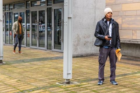 Young man standing at commercial entrance holding smartphone and mail envelope in urban plaza