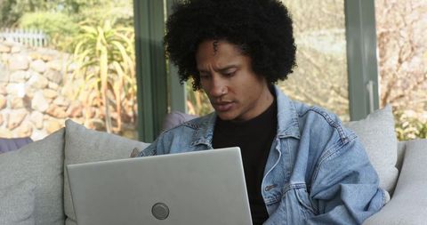 Working from home in sunroom, mid-adult man using laptop on gray sofa in denim jacket