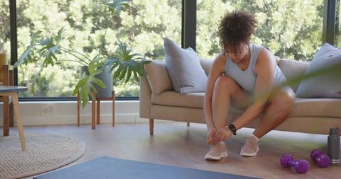 Woman tying shoelaces before home workout in sunlit living room with yoga mat and dumbbells