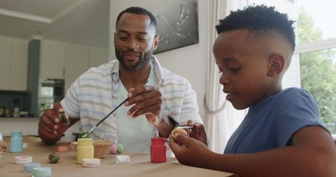 African american father and son painting easter eggs together in bright kitchen