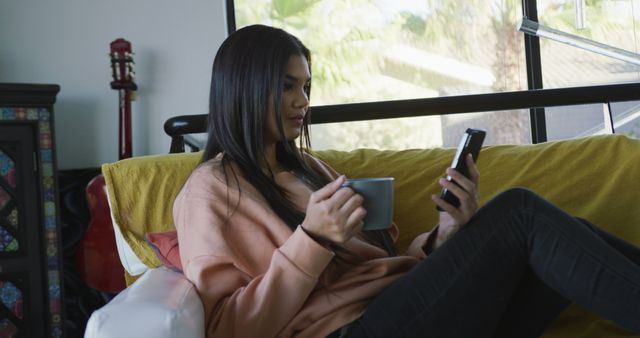 Image of biracial woman sitting on sofa, drinking coffee and using smartphone. leisure, relax, spending free time with technology at home concept.