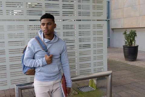 African american teen student standing by urban campus rail holding backpack and folder