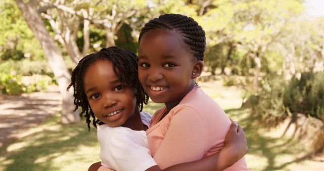 Two African American sisters smiling and hugging warmly outdoors in a park. Both kids are dressed casually and enjoying the sunny day surrounded by trees. This image is perfect for advertisements, social media posts, and websites promoting family values, outdoor activities, child-related content, and diversity representation.