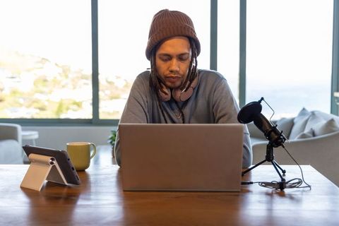 African american podcaster working remotely with laptop and microphone by coastal window