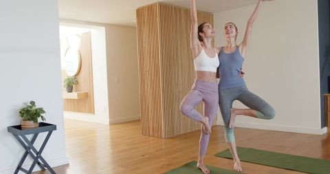 Two women practicing partner tree pose in minimalist home yoga studio