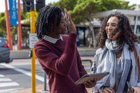 Young colleagues standing at crosswalk sharing tablet and laughing on sunny city street